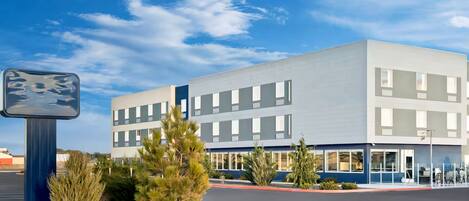 A modern building with a flat roof, rectangular windows, and a parking lot with a few trees and shrubs.