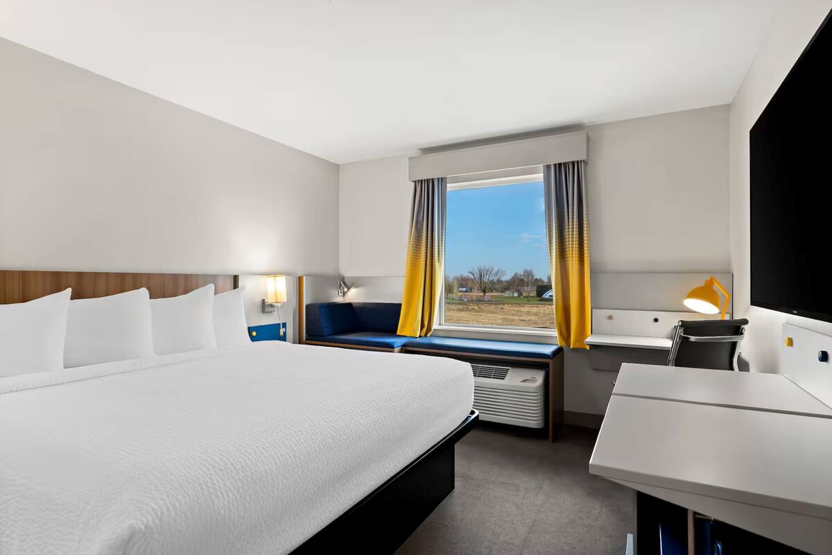 A hotel room with a large bed, a desk, a chair, a window with curtains, and a view of the outdoors.