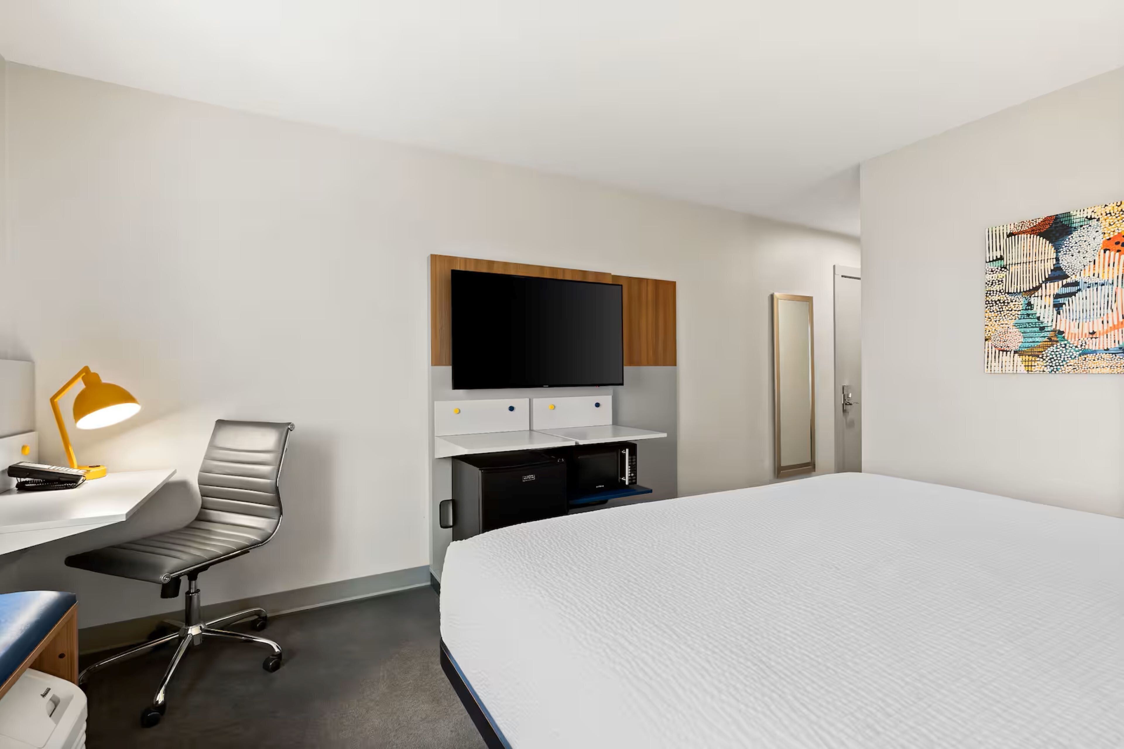 A hotel room with a bed, a desk with a chair, a TV mounted above the desk, and a door.