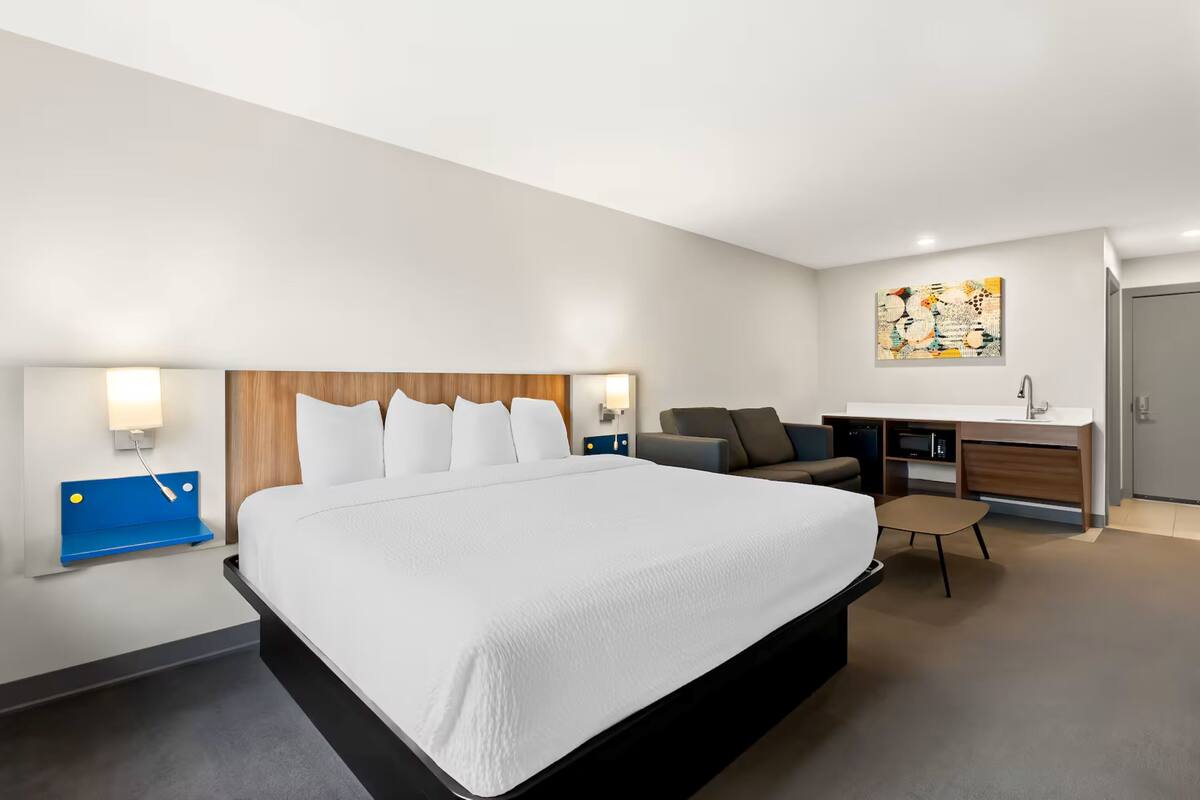 A modern hotel room with a large bed, a desk, a chair, a small table, and a bathroom with a sink.