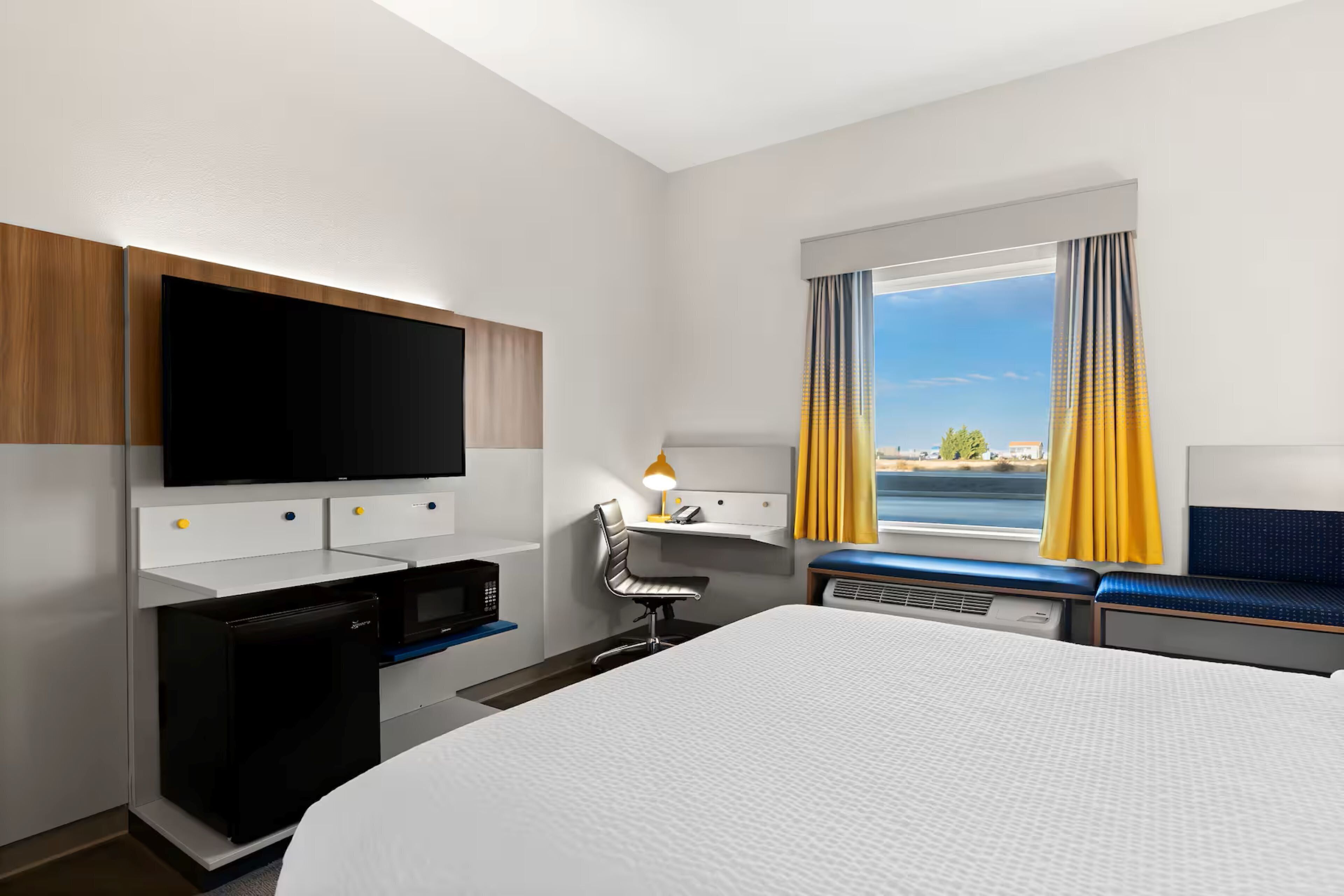 A hotel room with a bed, a desk with a computer, a TV, a small fridge, and a window with curtains.