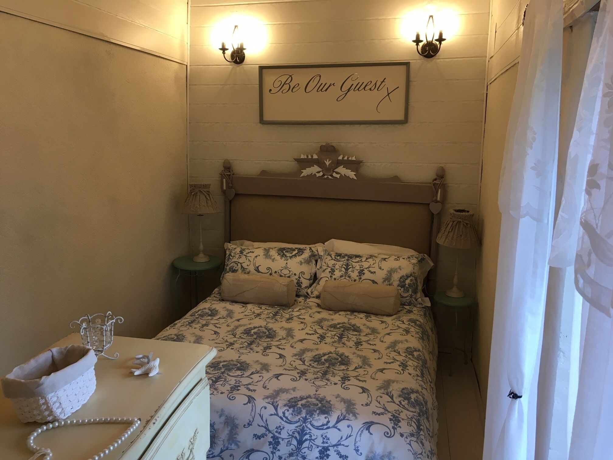 A neatly arranged bedroom with a bed, bedside tables, a lamp, a decorative sign, and a small dresser with a telephone and a decorative item.
