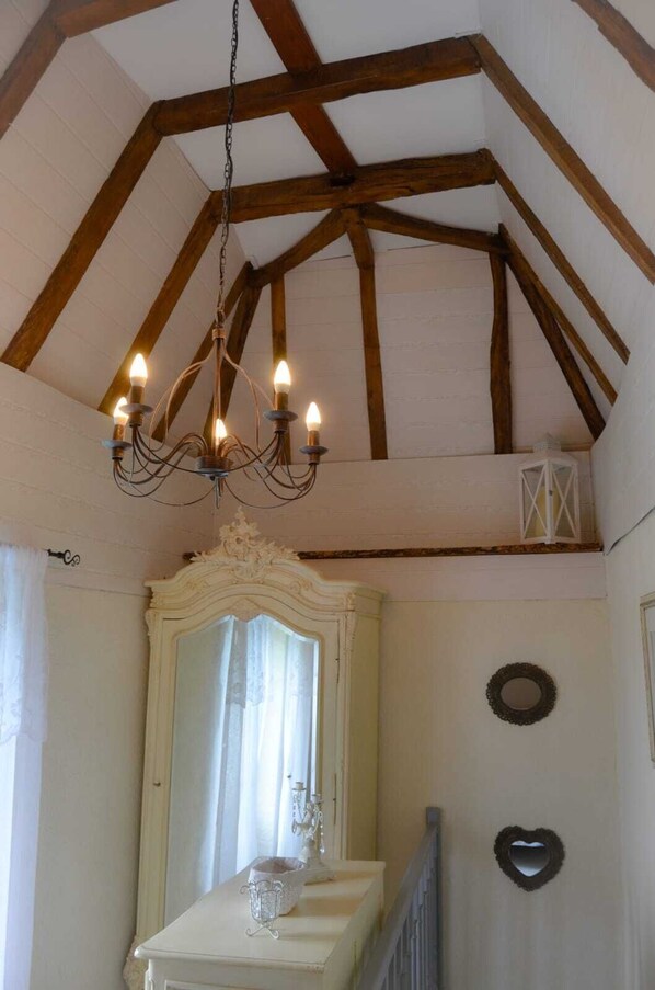 Una habitación con techo alto, vigas de madera a la vista, una araña, un espejo grande y ornamentado, y una pequeña mesa con una vela.