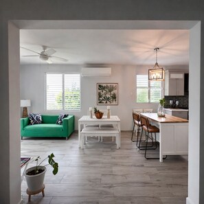 Un vaste espace de vie comprenant un canapé vert, une table à manger blanche et une cuisine dotée de meubles blancs et d’un plan de travail en bois.