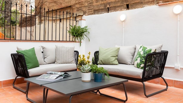 Ein gemütlicher Außenbereich mit einer Couch und Sesseln, einem Couchtisch mit Pflanzen und einer Ziegelwand im Hintergrund.
