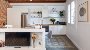 Eine moderne Küche mit weißen Schränken, einem Edelstahlkühlschrank und einem eingebauten Backofen. Ein Essbereich mit einem weißen Tisch und einem grauen Stuhl ist vorhanden. Die Küche verfügt über eine Backsteinwand und eine Holzbalkendecke.
