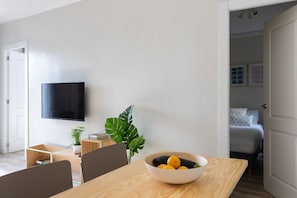 Ein modernes Wohnzimmer mit einem an der Wand befestigten Flachbildfernseher, einem Esstisch mit einer Obstschale und einer großen, grünen Pflanze.