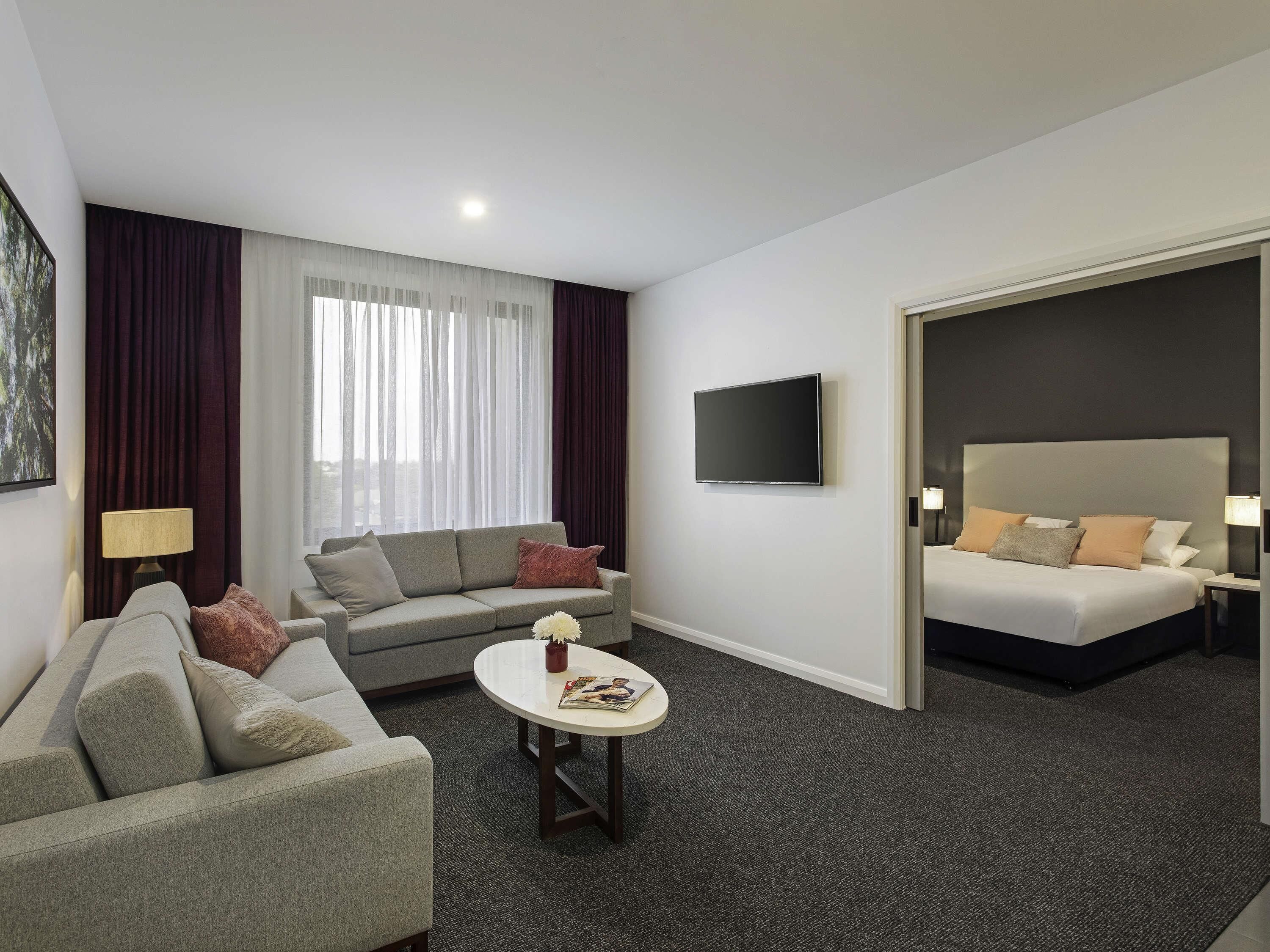 A hotel room with a sofa, a coffee table, a TV, a bed, and a view through a door.