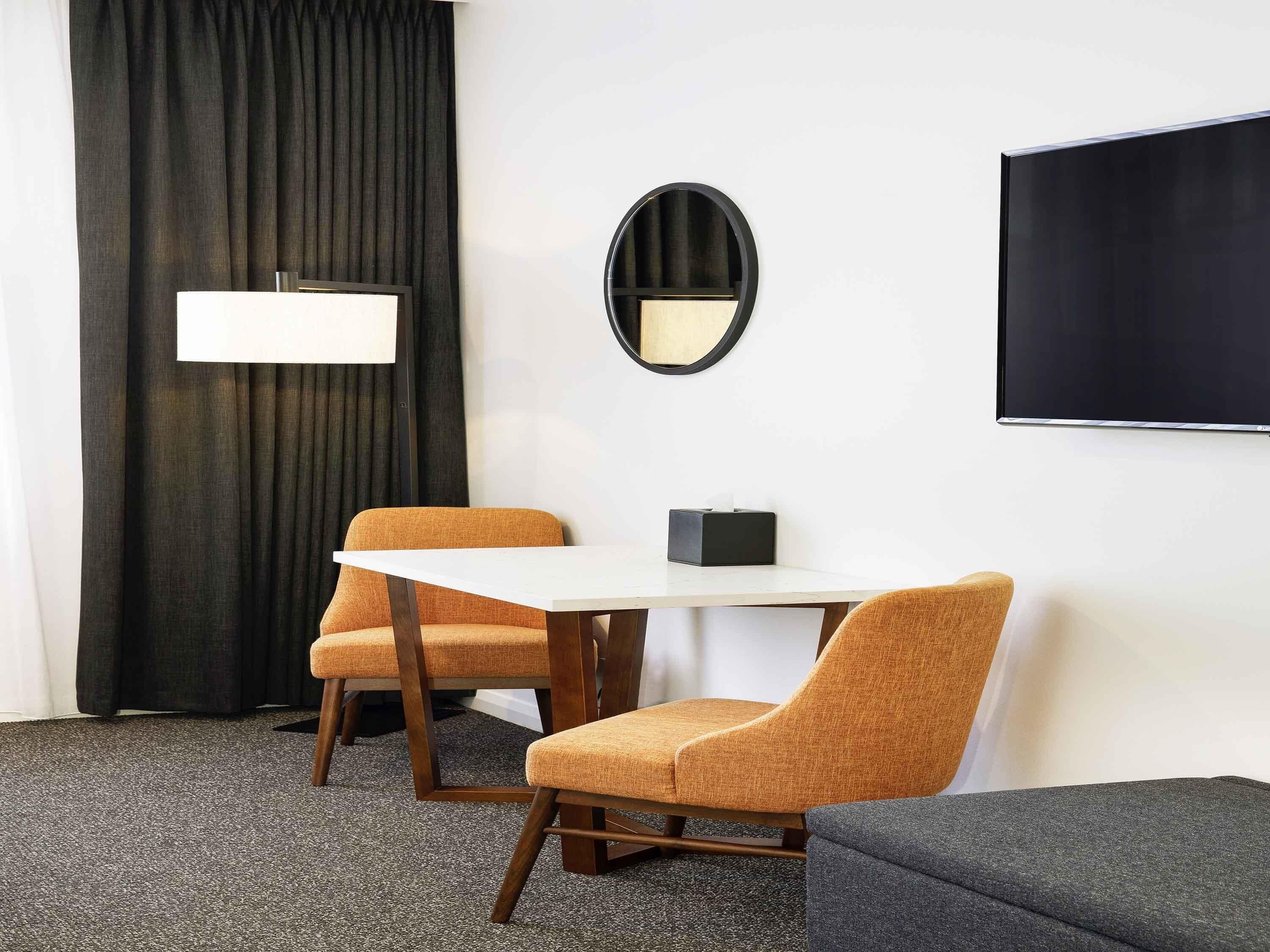A room with a white table, two orange chairs, a black-framed mirror, a black TV, and a black curtain.