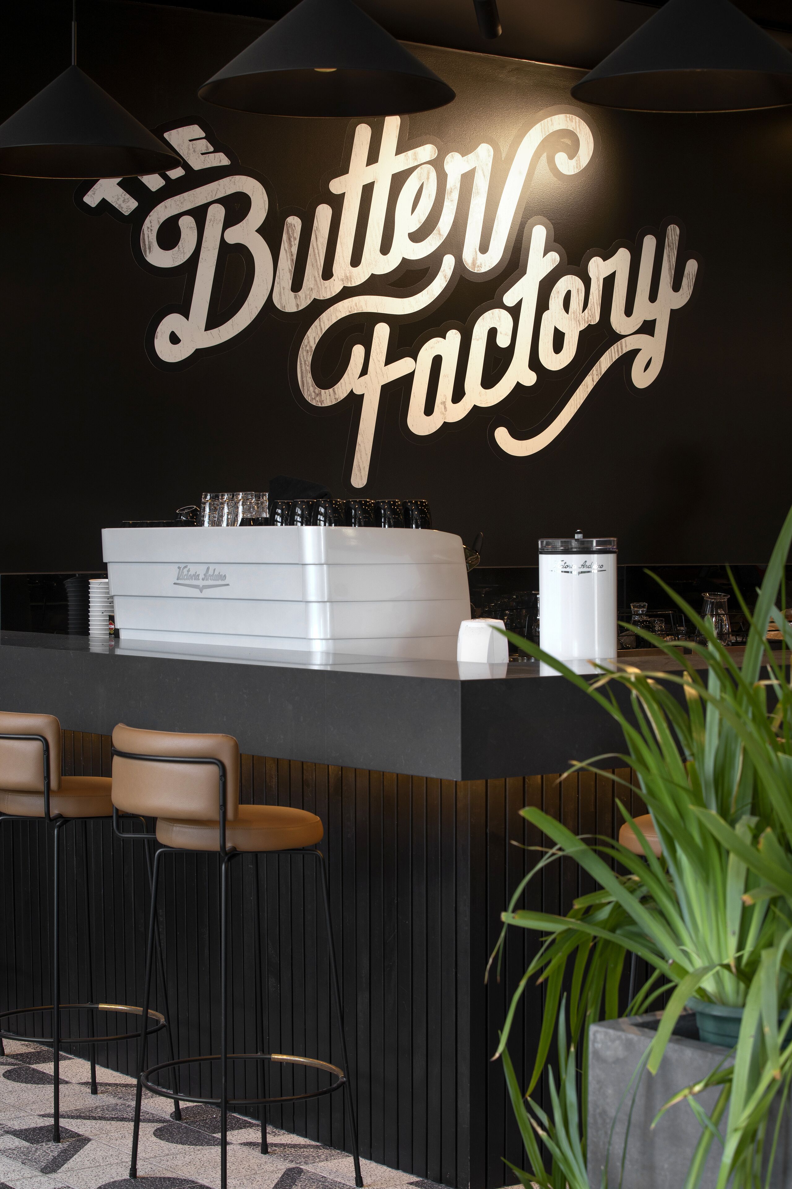 Uma área de bar com uma placa indicando "The Butter Factory", banquetas, uma máquina de café e um vaso de planta.