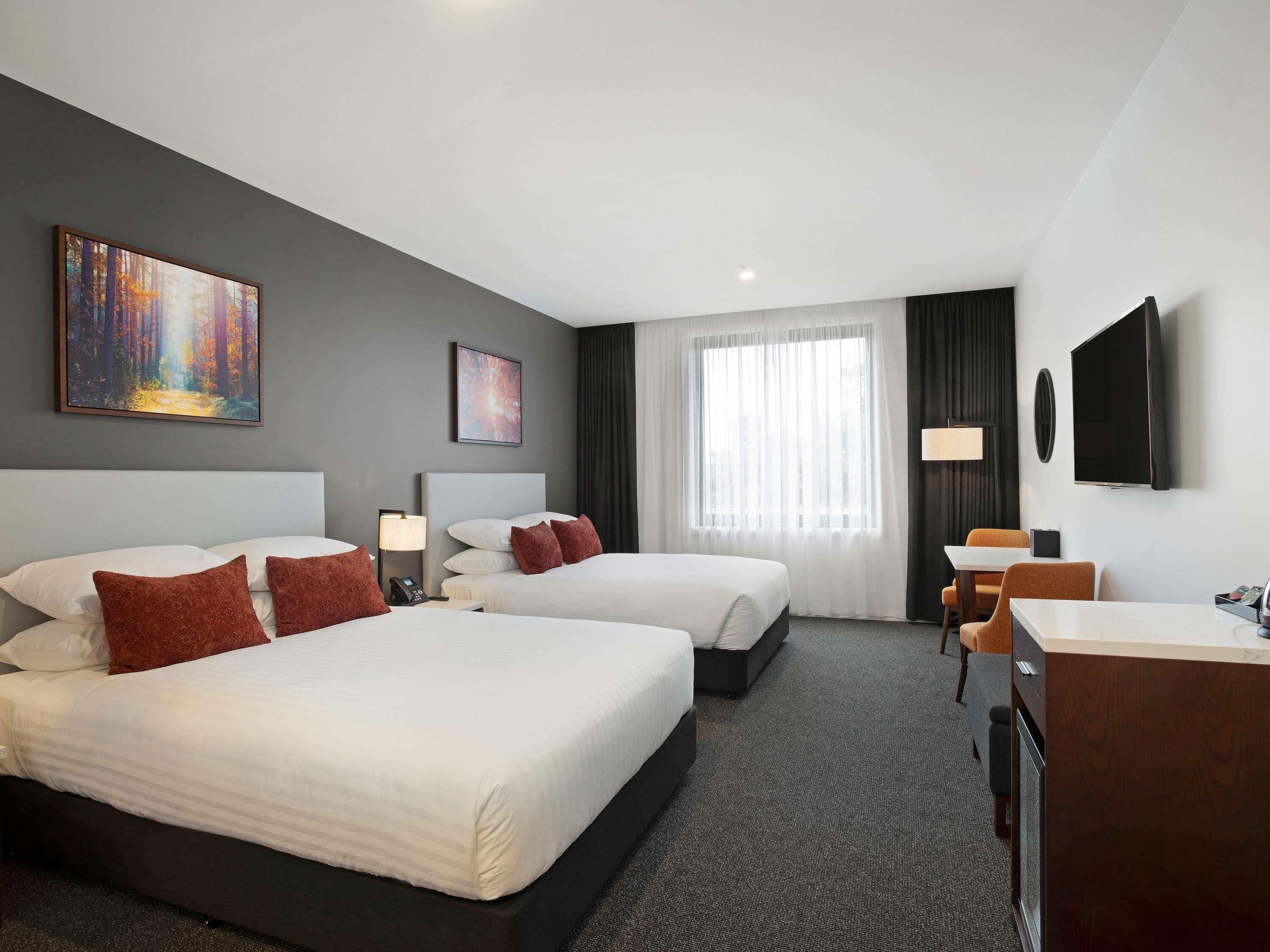 A hotel room with two beds, a desk, a chair, a TV, and artwork on the walls.