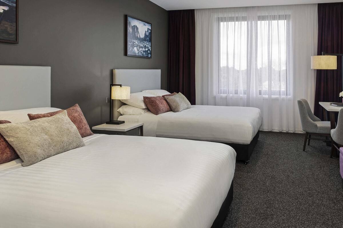 Habitación de hotel con dos camas, un escritorio, una silla y un ventanal grande con cortinas traslúcidas.