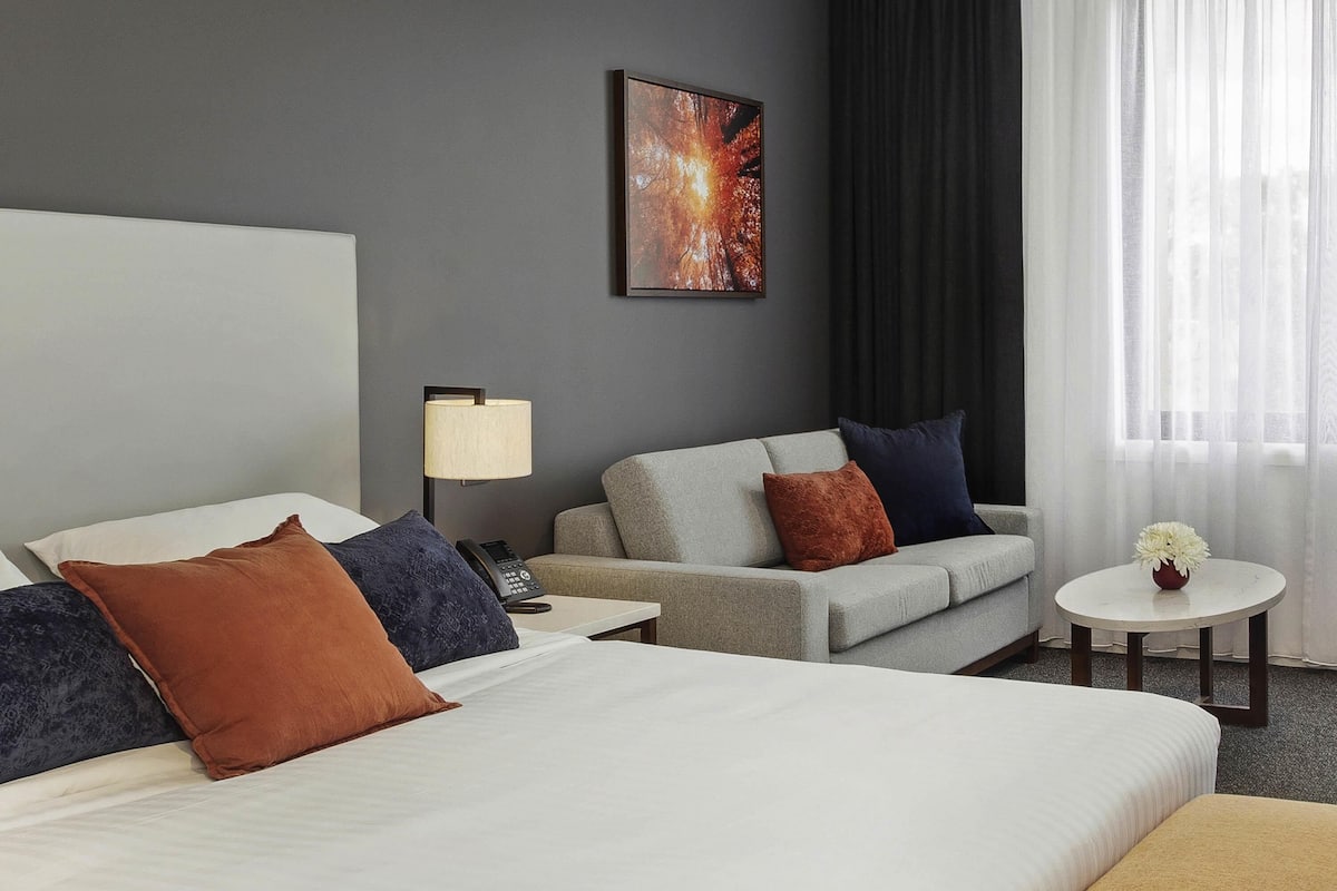 Habitación de hotel con cama, sofá, una mesita con un jarrón, lámpara y un cuadro en la pared.