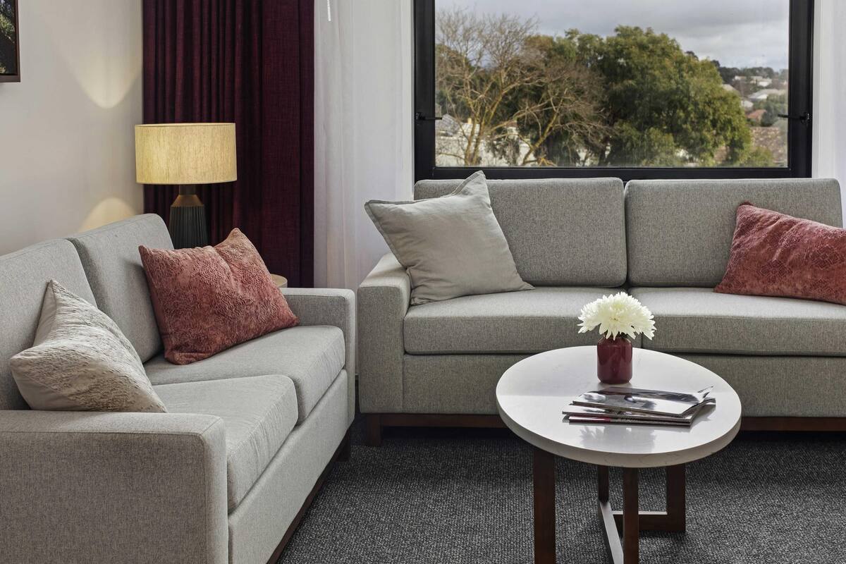 Un salón moderno con un sofá gris, una mesita redonda y vistas a árboles y casas a través de una ventana.