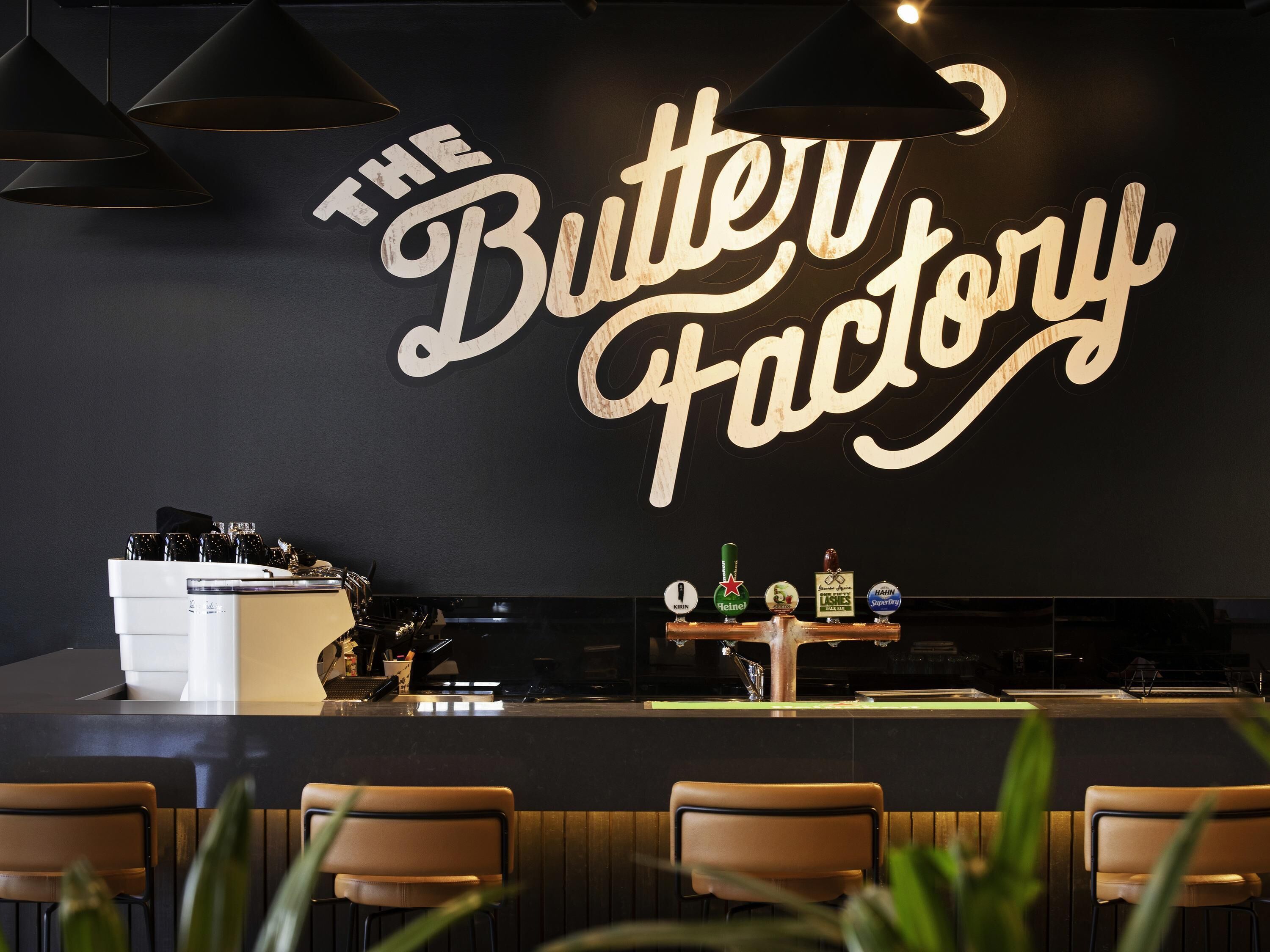 Een bar met een bord waarop 'The Butter Factory' staat, barstoelen, een toonbank met een koffieapparaat en verschillende flessen op de plank.