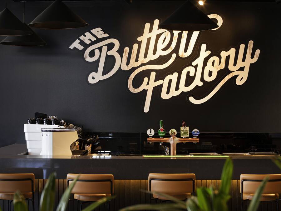 Een bar met een bord waarop 'The Butter Factory' staat, barstoelen, een toonbank met een koffieapparaat en verschillende flessen op de plank.