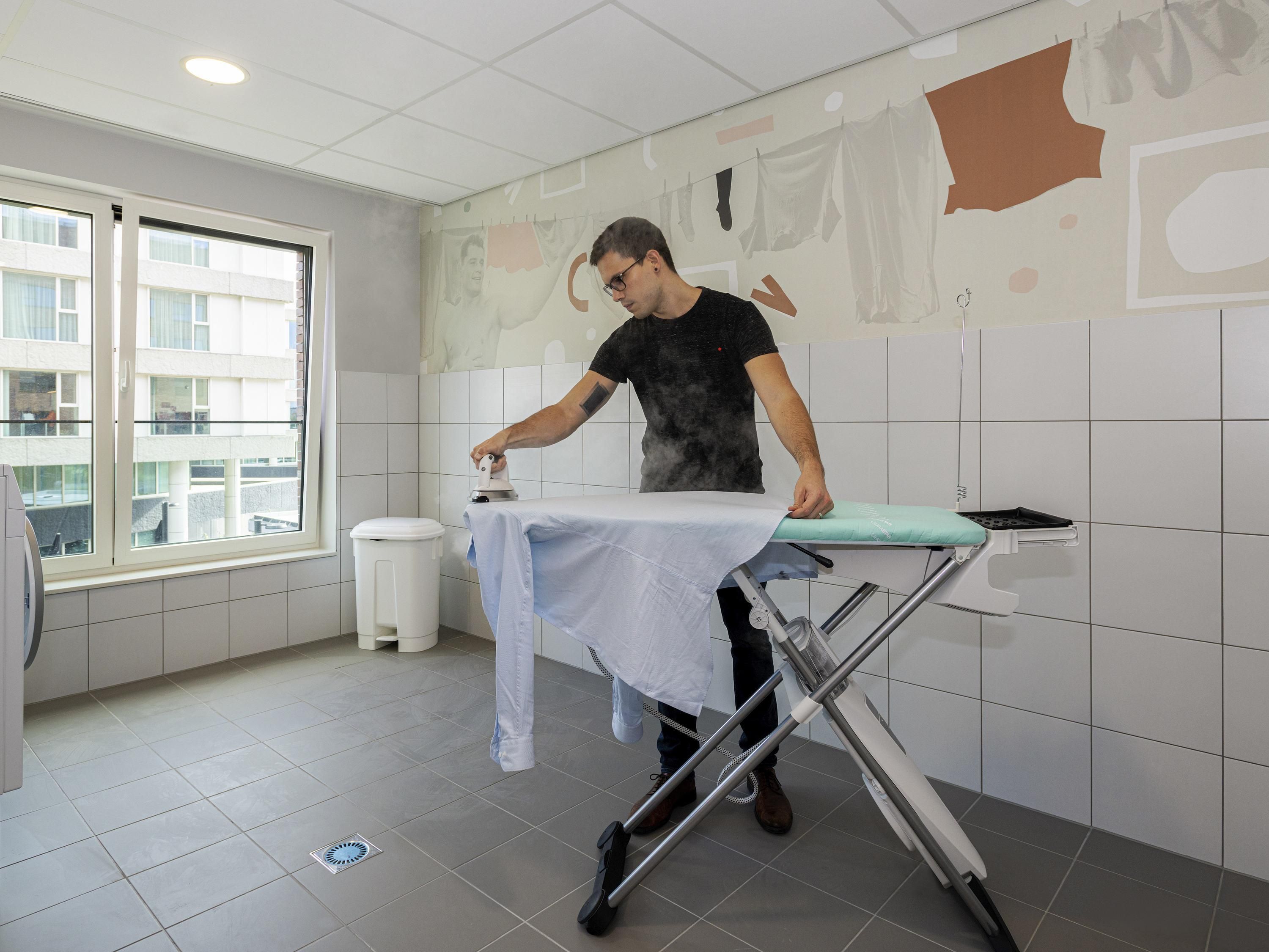 Una persona planchando una camisa blanca sobre una tabla de planchar en una habitación con paredes de azulejos blancos y una ventana.