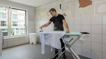 Una persona planchando una camisa blanca sobre una tabla de planchar en una habitación con paredes de azulejos blancos y una ventana.