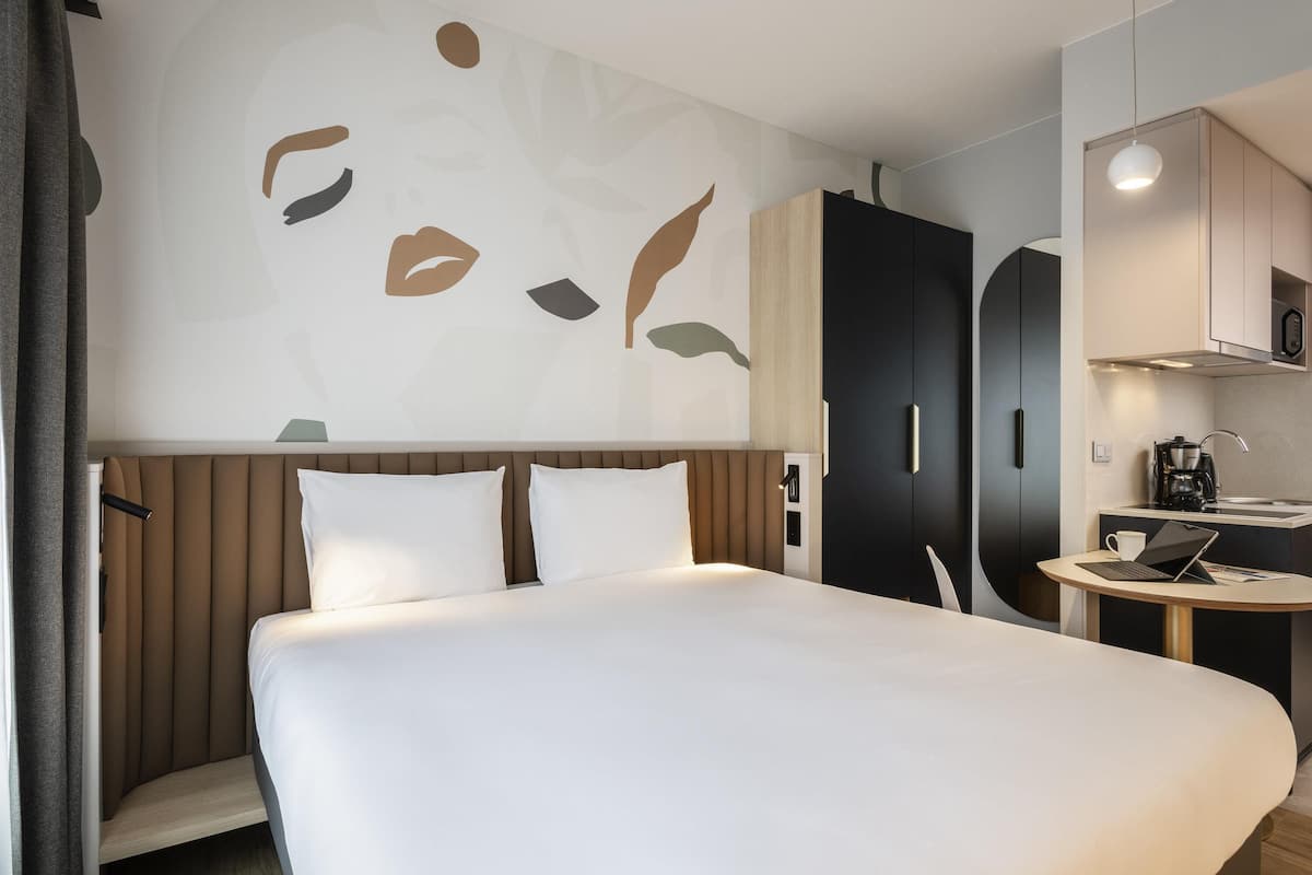 A hotel room with a large bed, a desk with a laptop, a wardrobe, and a kitchenette.
