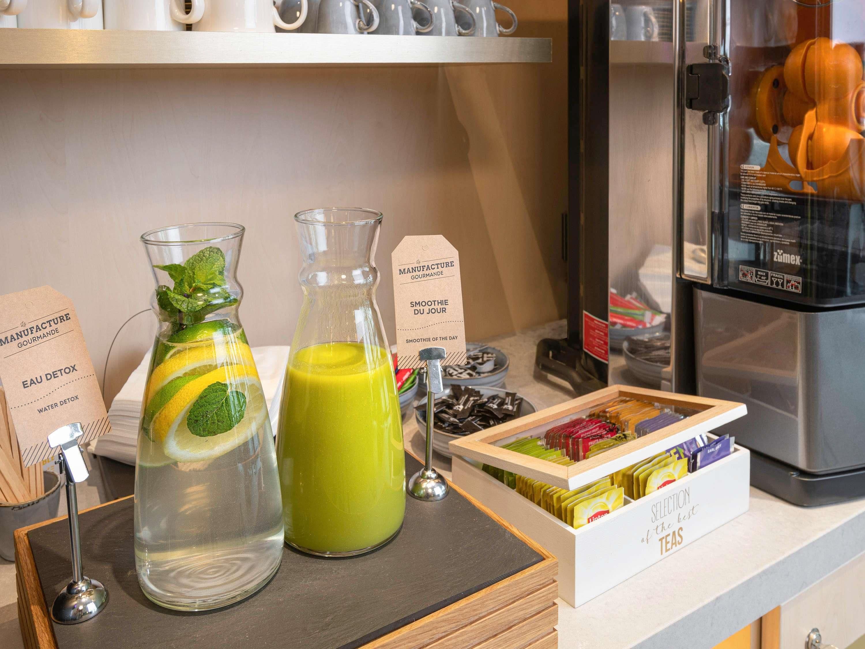 Una estación de bebidas con dos jarras, una con una bebida verde y otra con agua, además de una caja de bolsitas de té.
