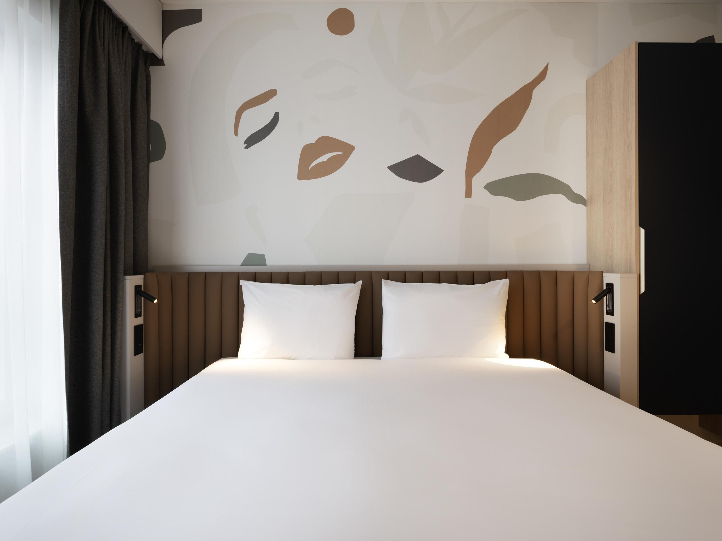 A hotel room with a bed, two pillows, a headboard, and a wall with abstract art.