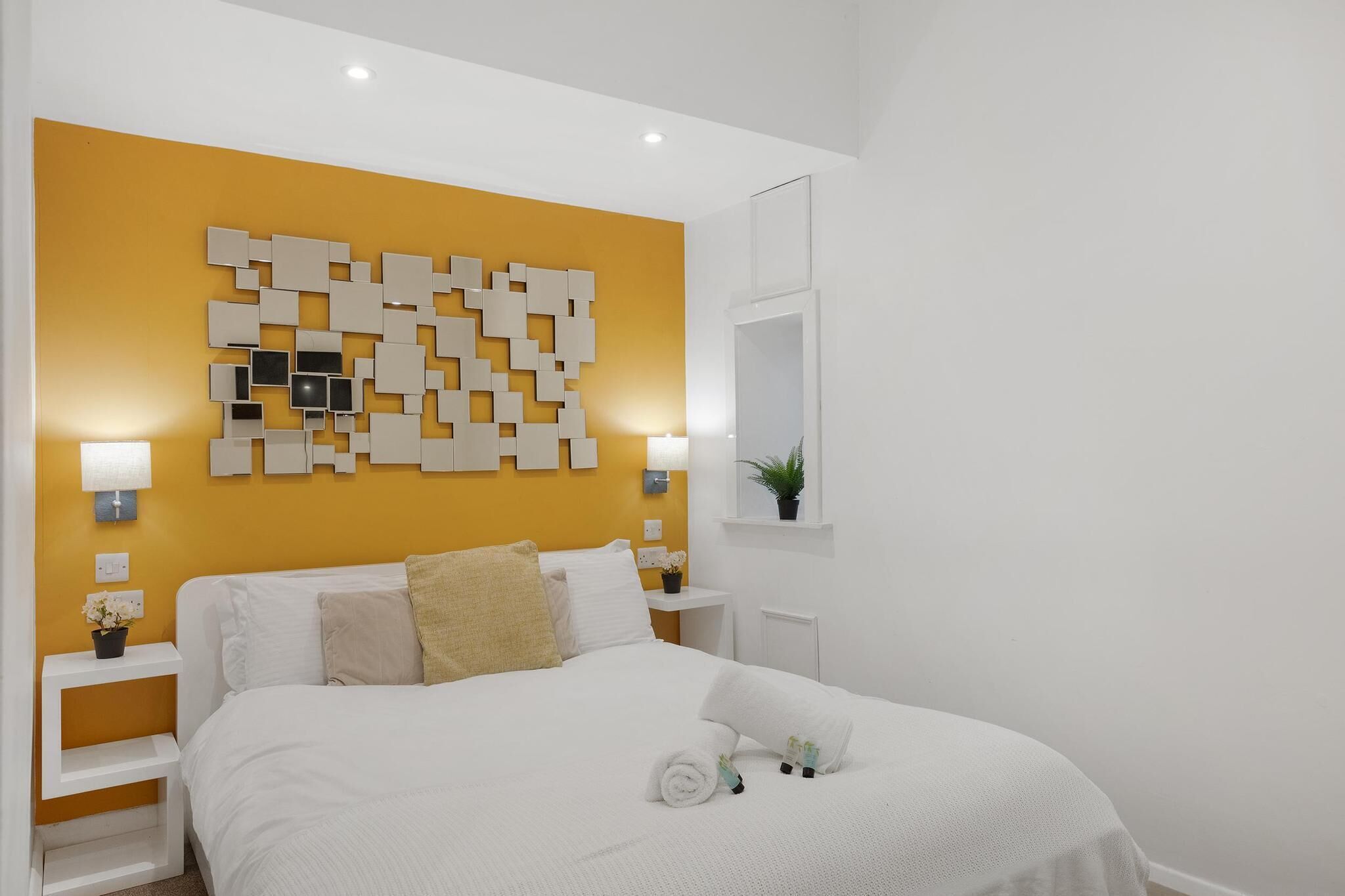 Ett modernt hotellrum med en stor säng, en dekorativ väggpanel, sänglampor, ett litet bord med en växt och en spegel.