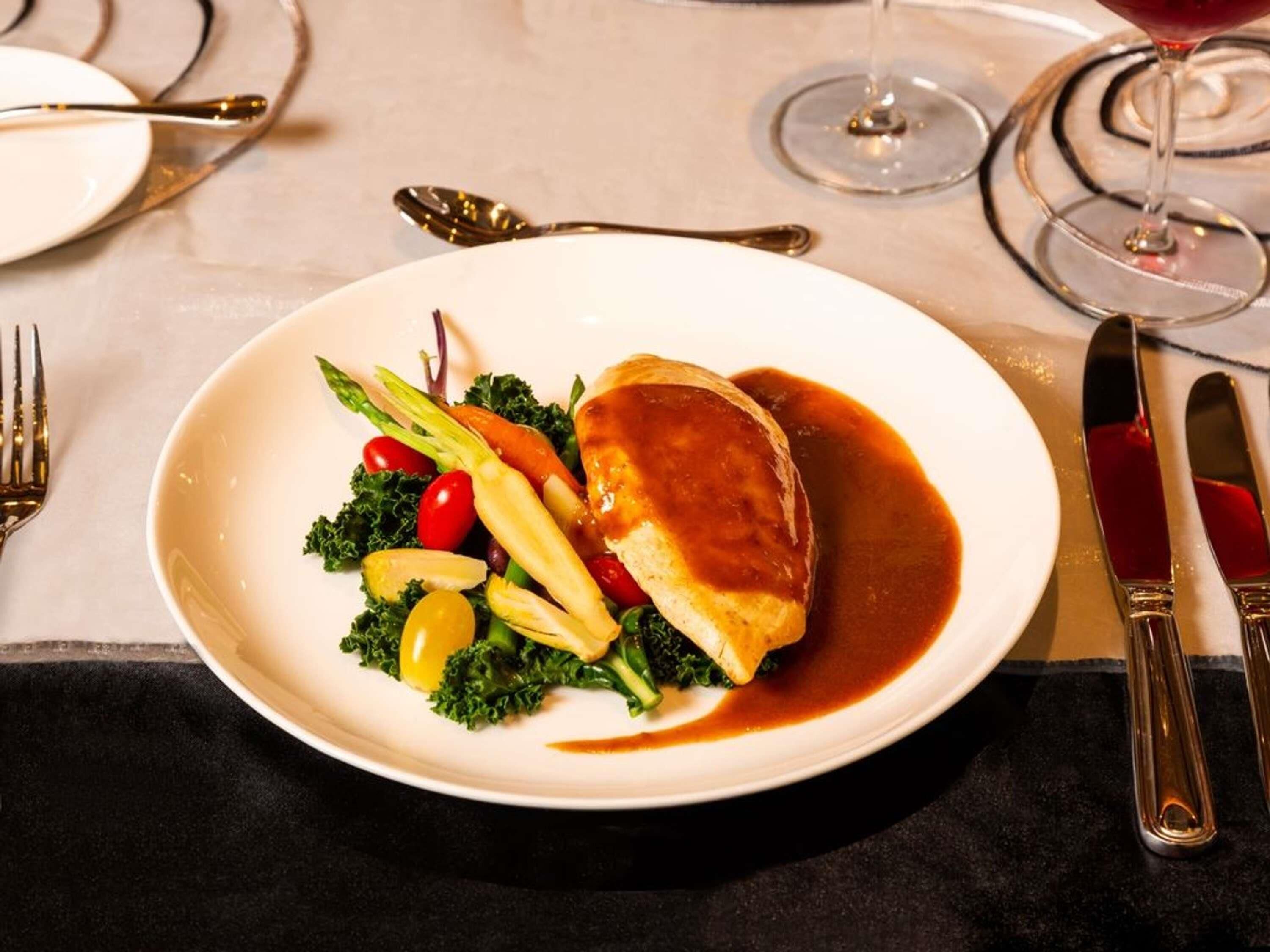 A plate with a meat dish, vegetables, and sauce on a table with cutlery and a glass of red wine.