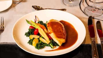 A plate with a meat dish, vegetables, and sauce on a table with cutlery and a glass of red wine.