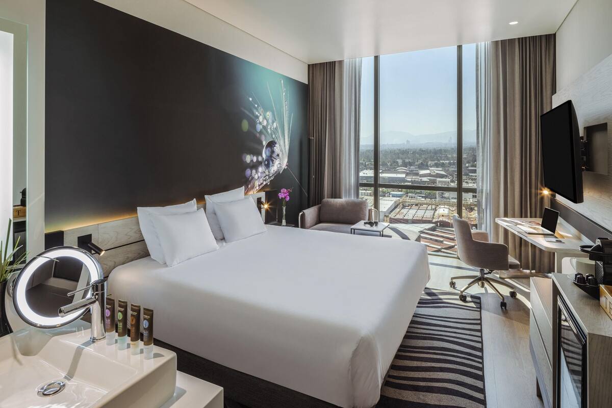 A modern hotel room with a large bed, a desk with a laptop, a TV, and a view of a cityscape.