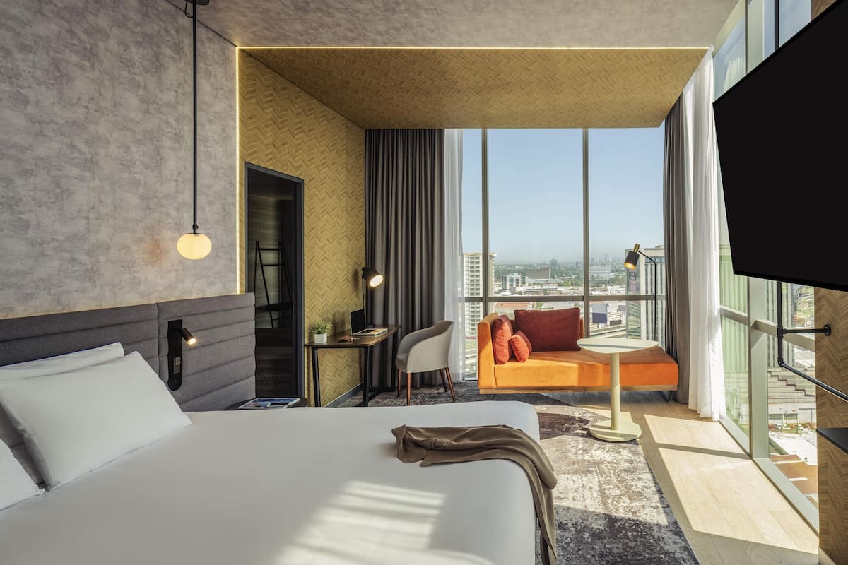 A modern hotel room with a large bed, a desk, a chair, and a sofa. There is a TV mounted on the wall and a window with a city view.