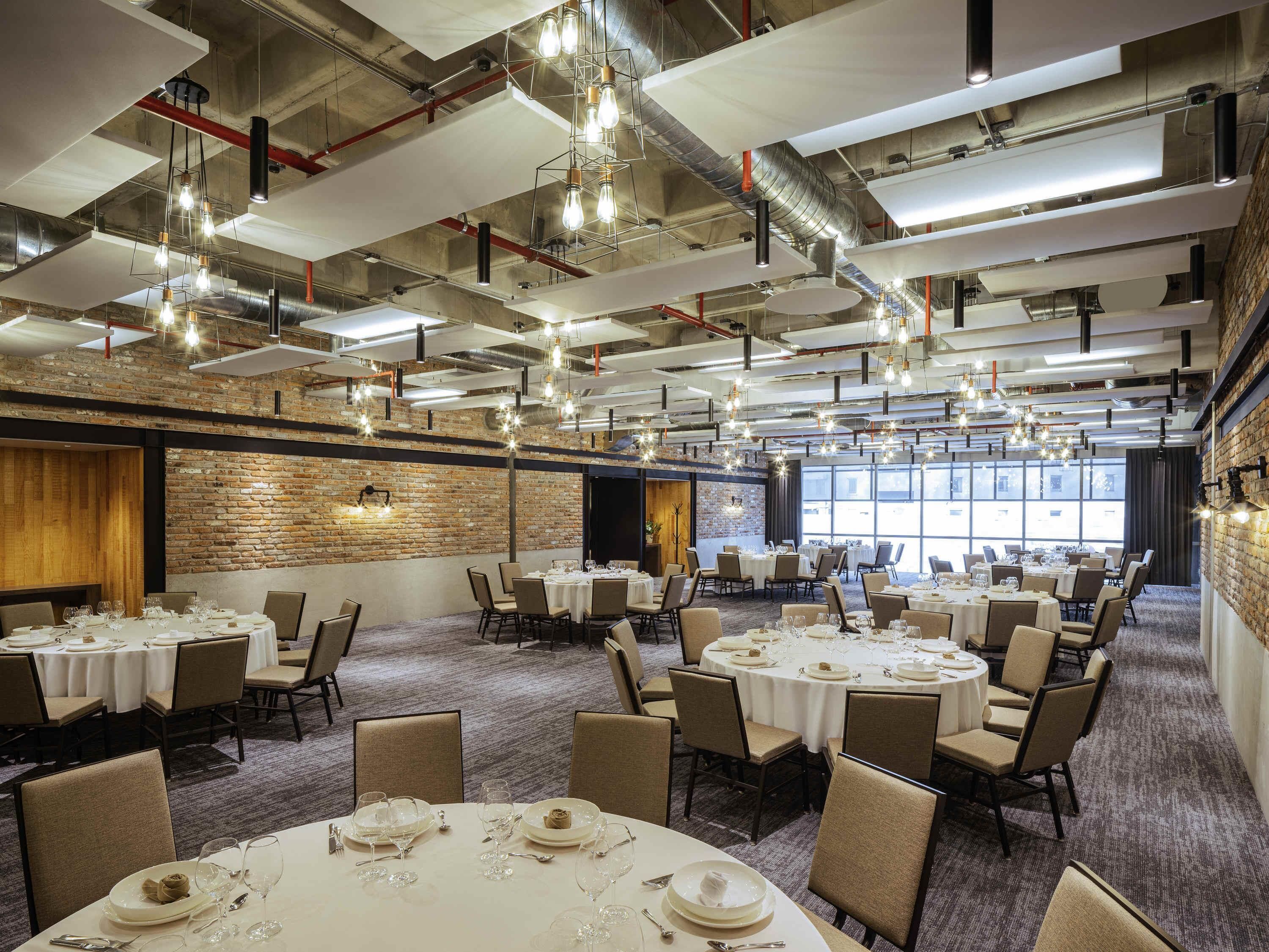 A spacious banquet hall with multiple round tables set for dining, exposed ductwork, and a brick wall.
