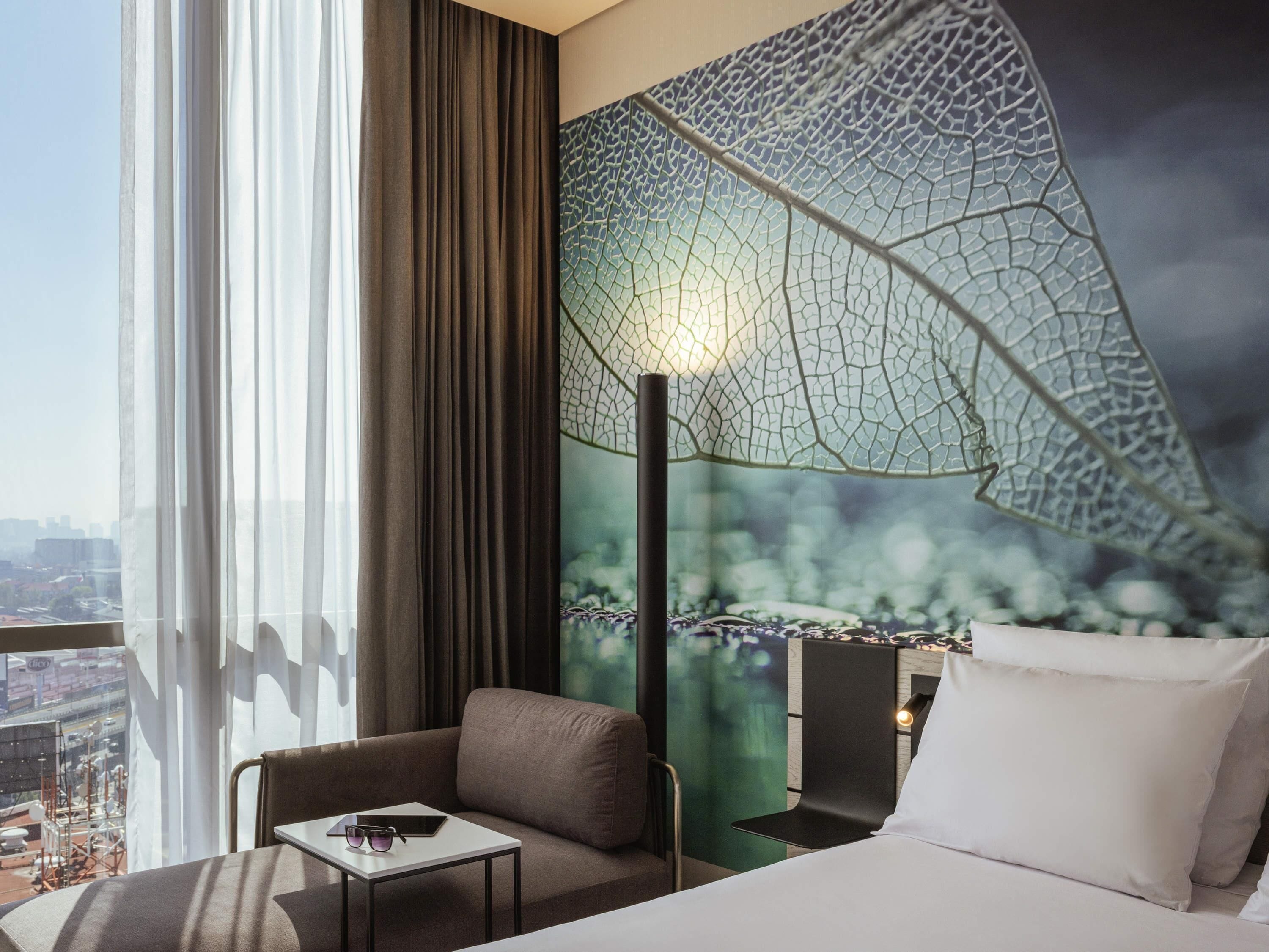 A hotel room with a bed, a chair, a small table, a large window with curtains, and a wall mural of a dragonfly's wing.