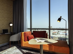 Una habitación de hotel moderna con un ventanal grande, un sofá, una mesita con un libro y vasos, y un escritorio con una computadora portátil.