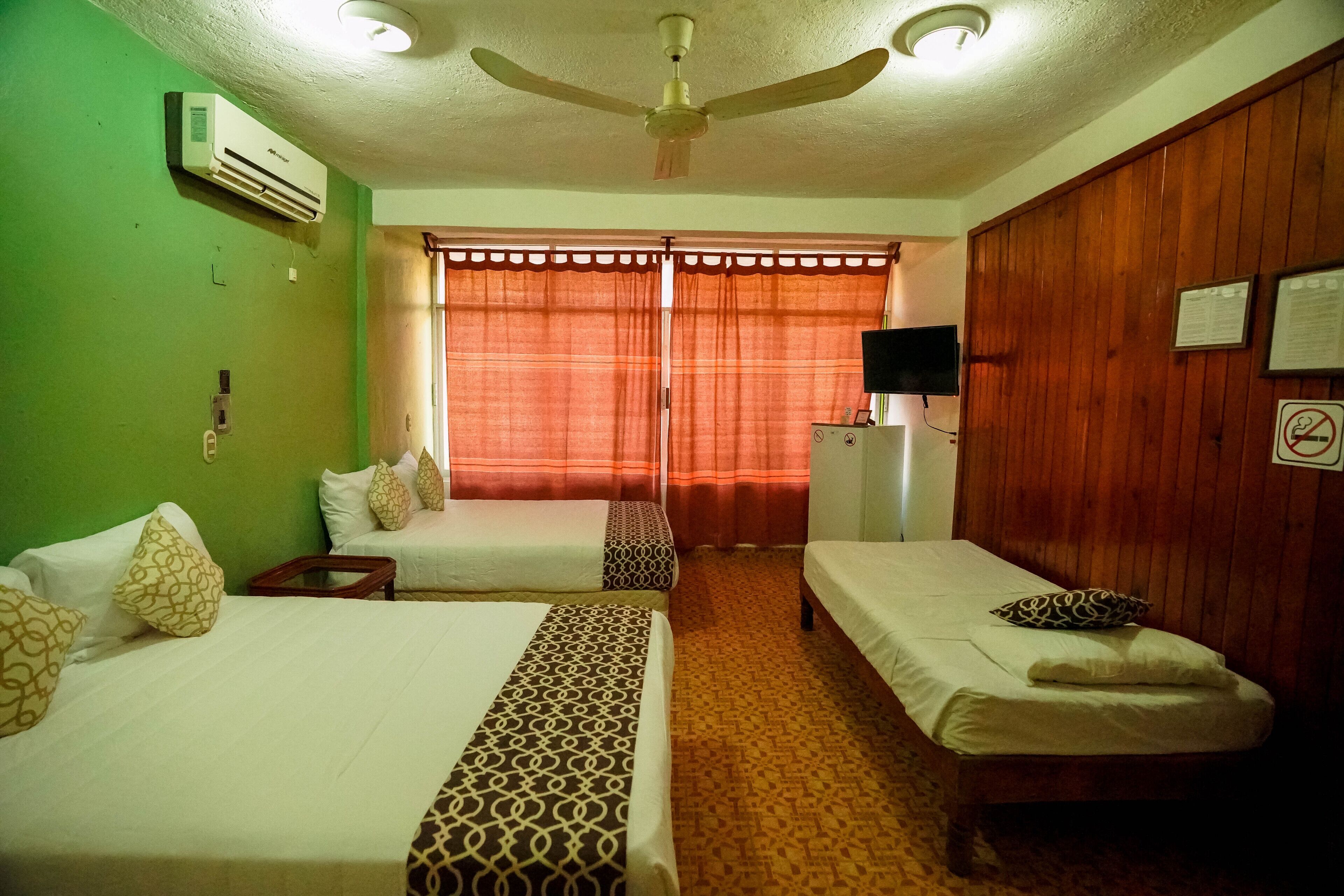 A hotel room with two single beds, a ceiling fan, an air conditioner, a television on a wooden cabinet, and a window with curtains.