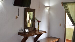 Una habitación con una televisión montada en la pared, una mesita consola de madera con un espejo, un jarrón con flores y un banco.