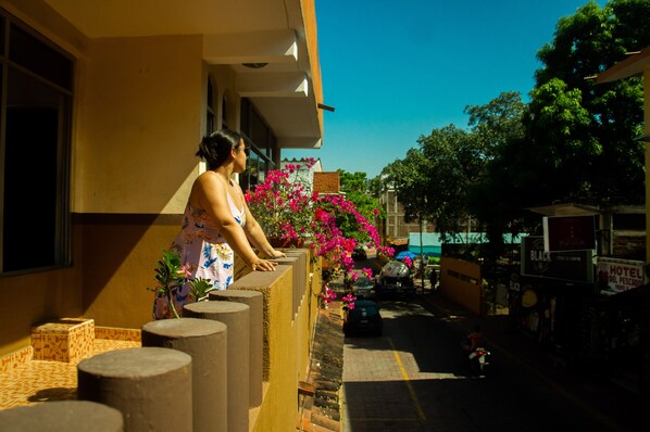 A woman standing on a balcony overlooking a street with a building, a parked car, and a motorcycle. - Hotel Casa Arena (Zihuatanejo)