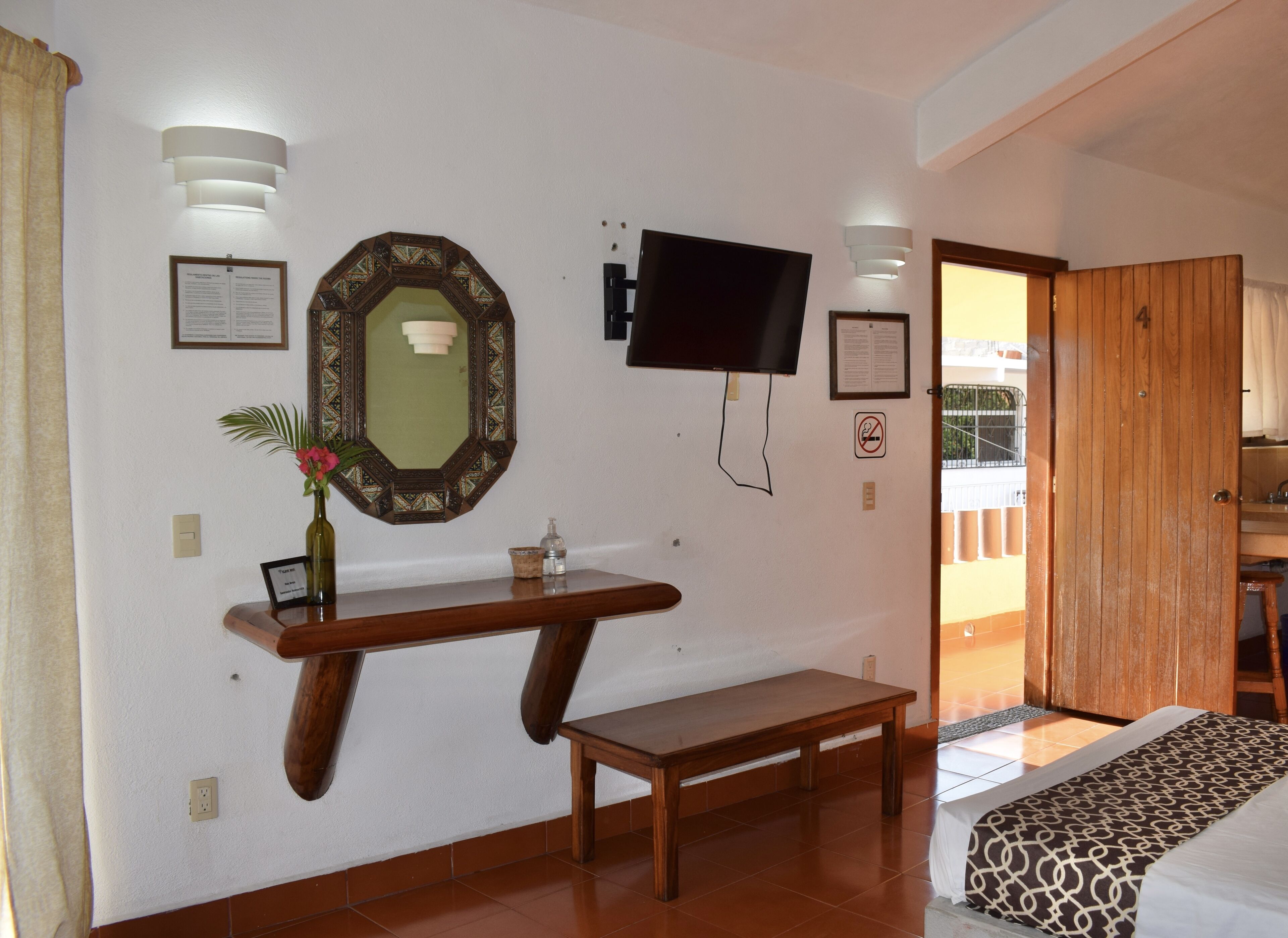 Quarto de hotel com um banco de madeira, televisão presa na parede, espelho com moldura decorativa e porta de madeira.