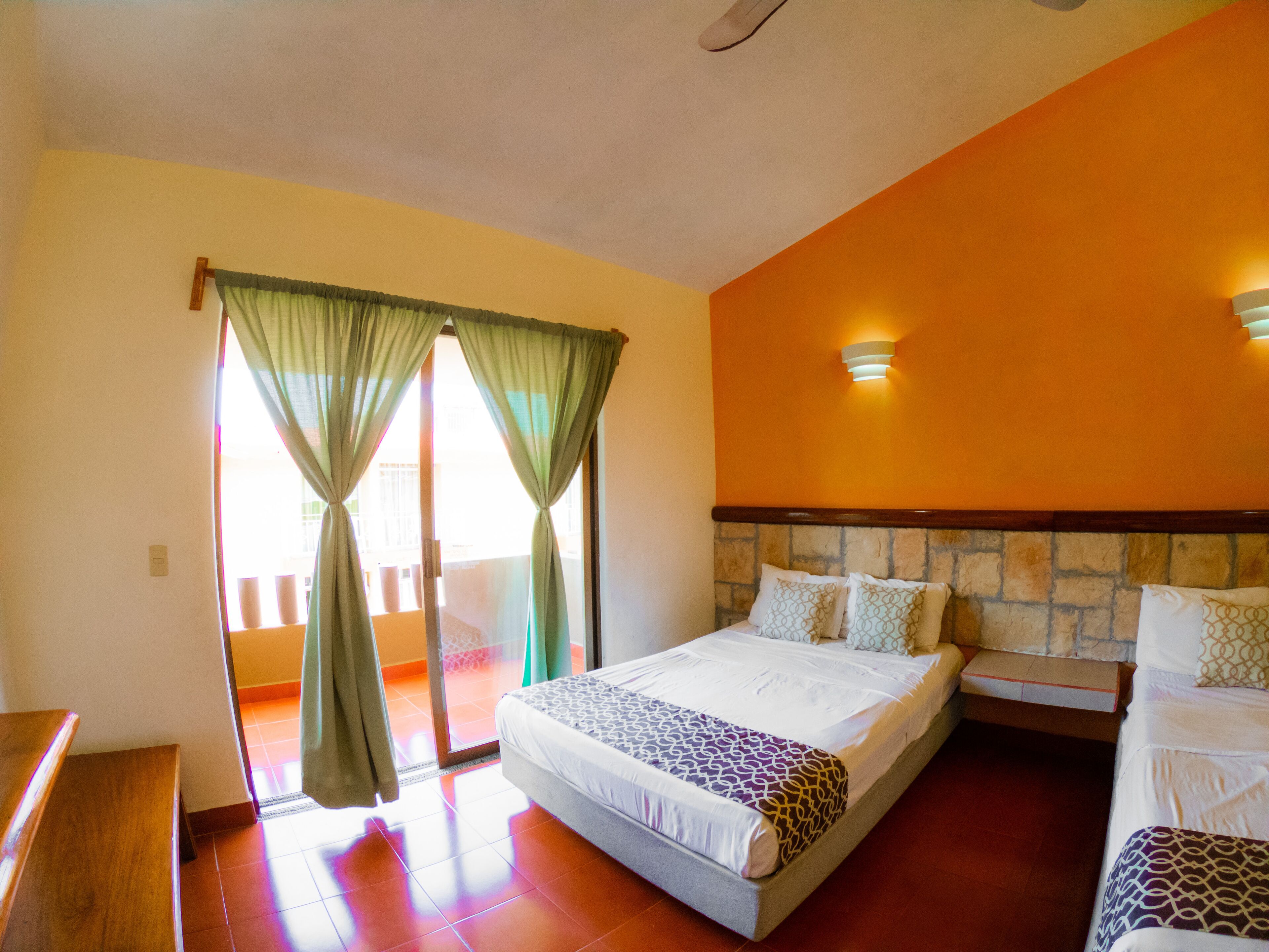 A hotel room with a large bed, a smaller bed, a wooden bench, a sliding glass door with curtains, and a stone accent wall.