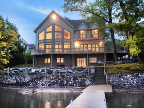 A large, multi-story house with a dock extending into a body of water, surrounded by a stone wall and trees.