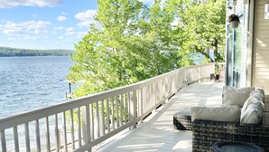 A wooden deck with a white railing overlooking a lake, a wicker sofa with cushions, and a hanging planter.