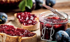 Un tarro de mermelada con una cuchara, una rebanada de pan y ciruelas enteras.