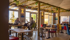 Un café avec des tables et des chaises en bois, de grandes fenêtres et des plantes suspendues.