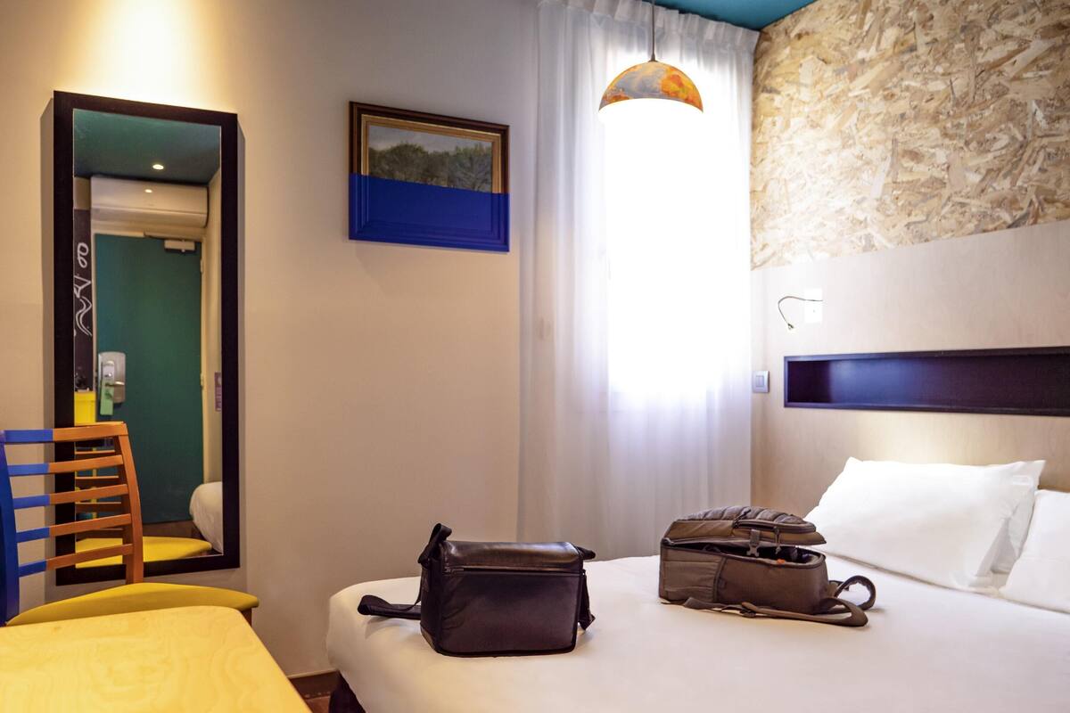 Habitación de hotel con cama, escritorio, silla, lámpara de escritorio, ventana con cortinas y un cuadro en la pared.