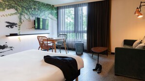 A hotel room with a bed, a desk with chairs, a television, a window with curtains, and a sofa.