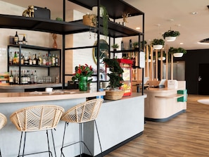 Zona de bar con mostrador, taburetes, estanterías con botellas y plantas decorativas.