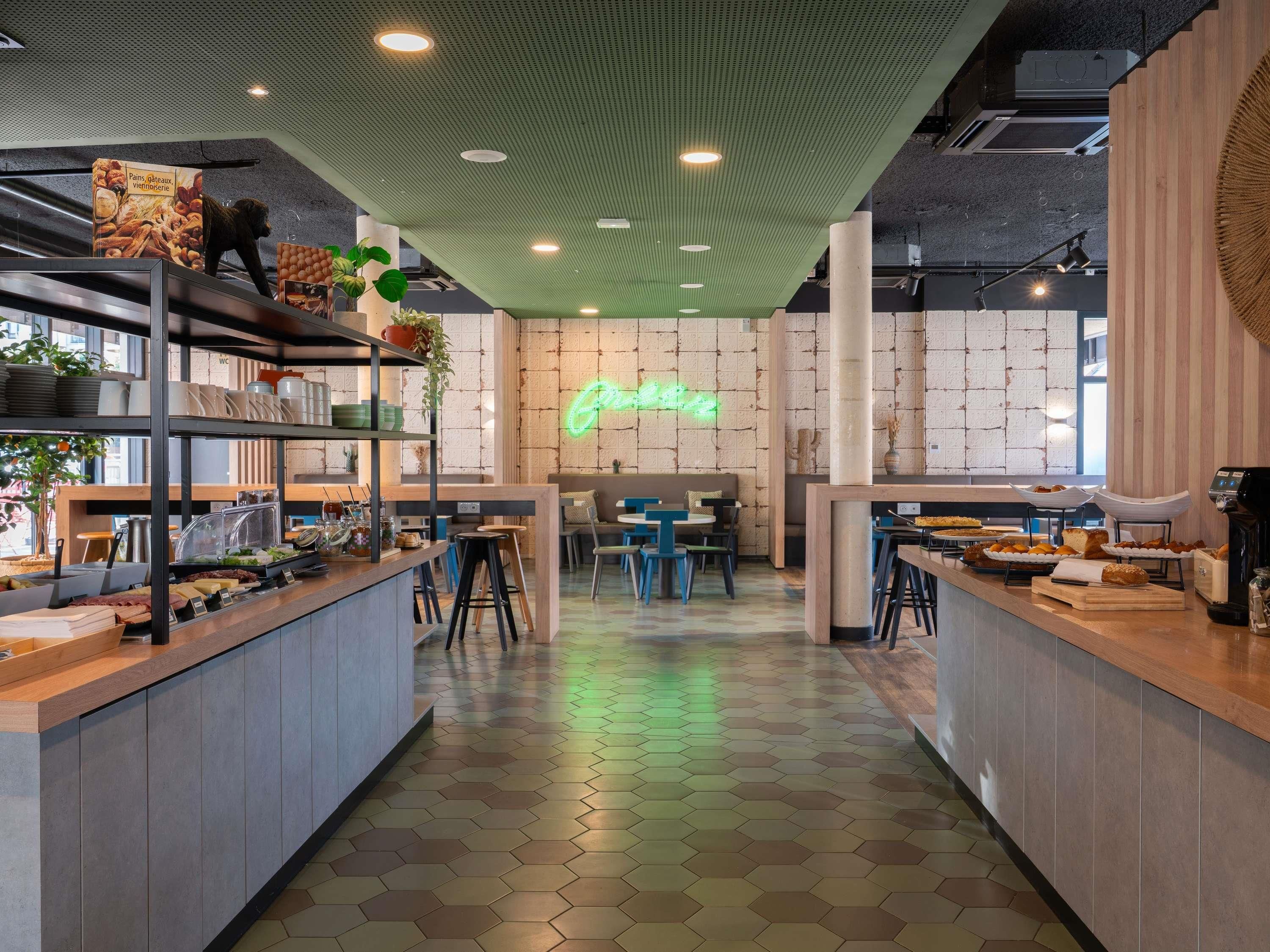 Interior de una cafetería con suelo de patrón hexagonal, mostrador de madera con exhibición de comida, zona de estar y un letrero de neón.