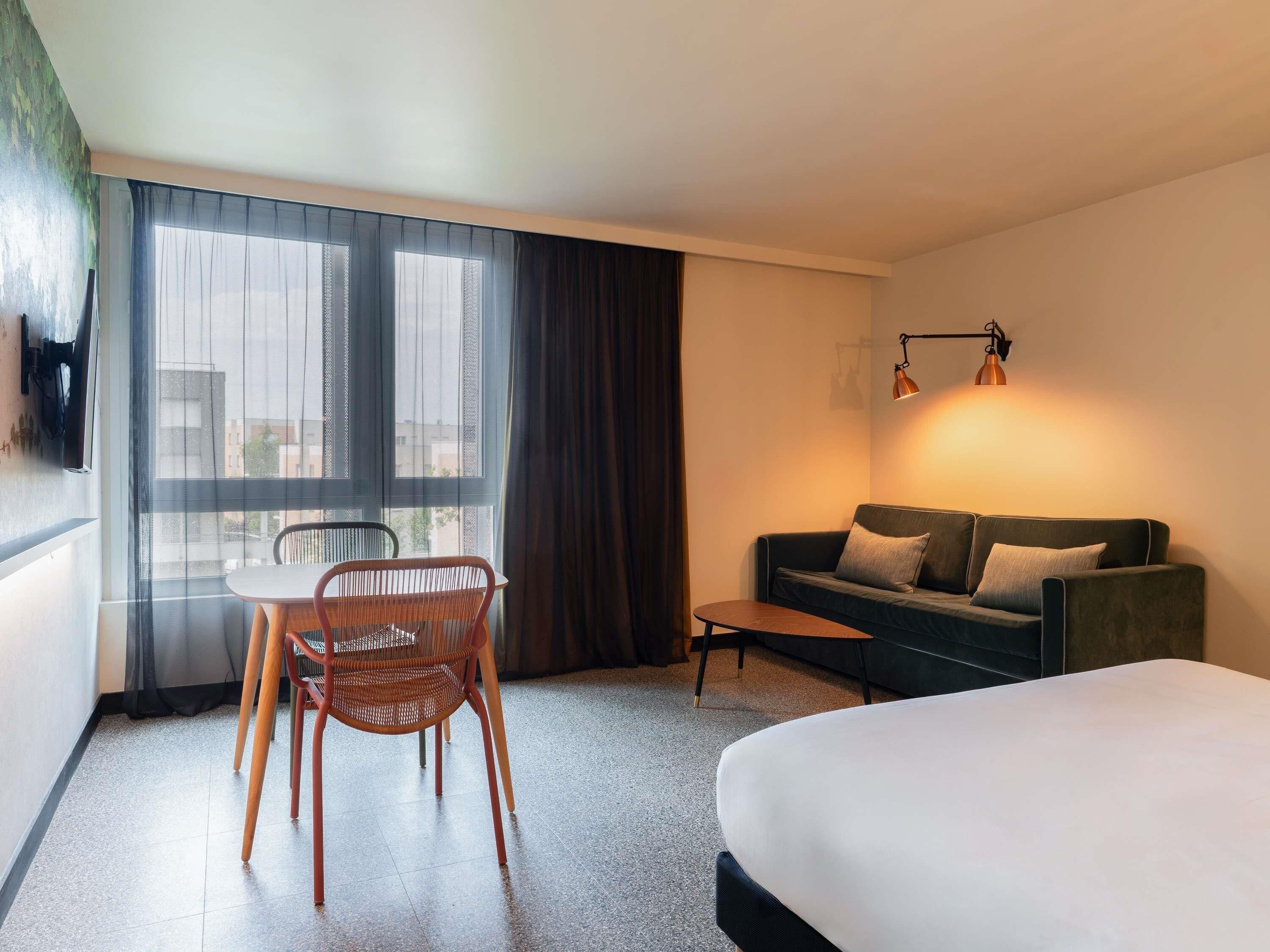 A hotel room with a bed, a sofa, a dining table with chairs, and a large window with curtains.
