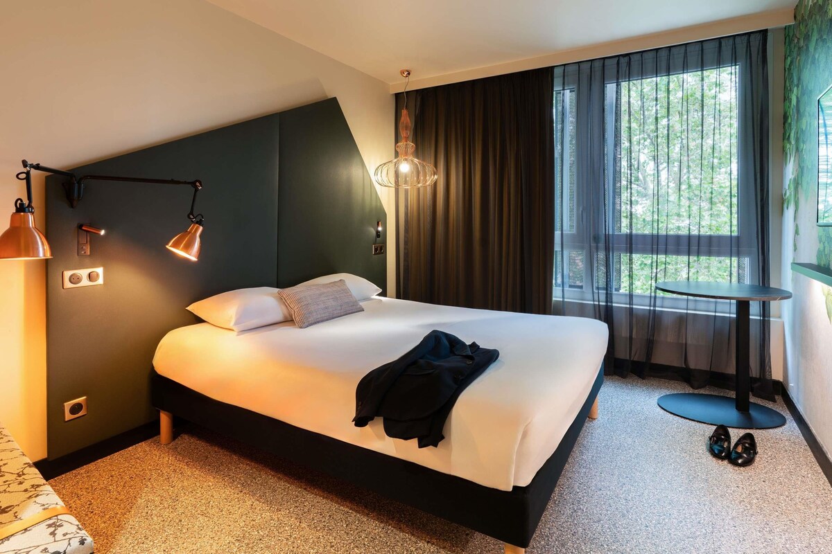 Habitación de hotel moderna con una cama grande, una mesita y vistas a zonas verdes por la ventana.