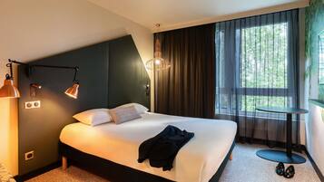 A modern hotel room with a large bed, a small table, and a view of greenery through the window.