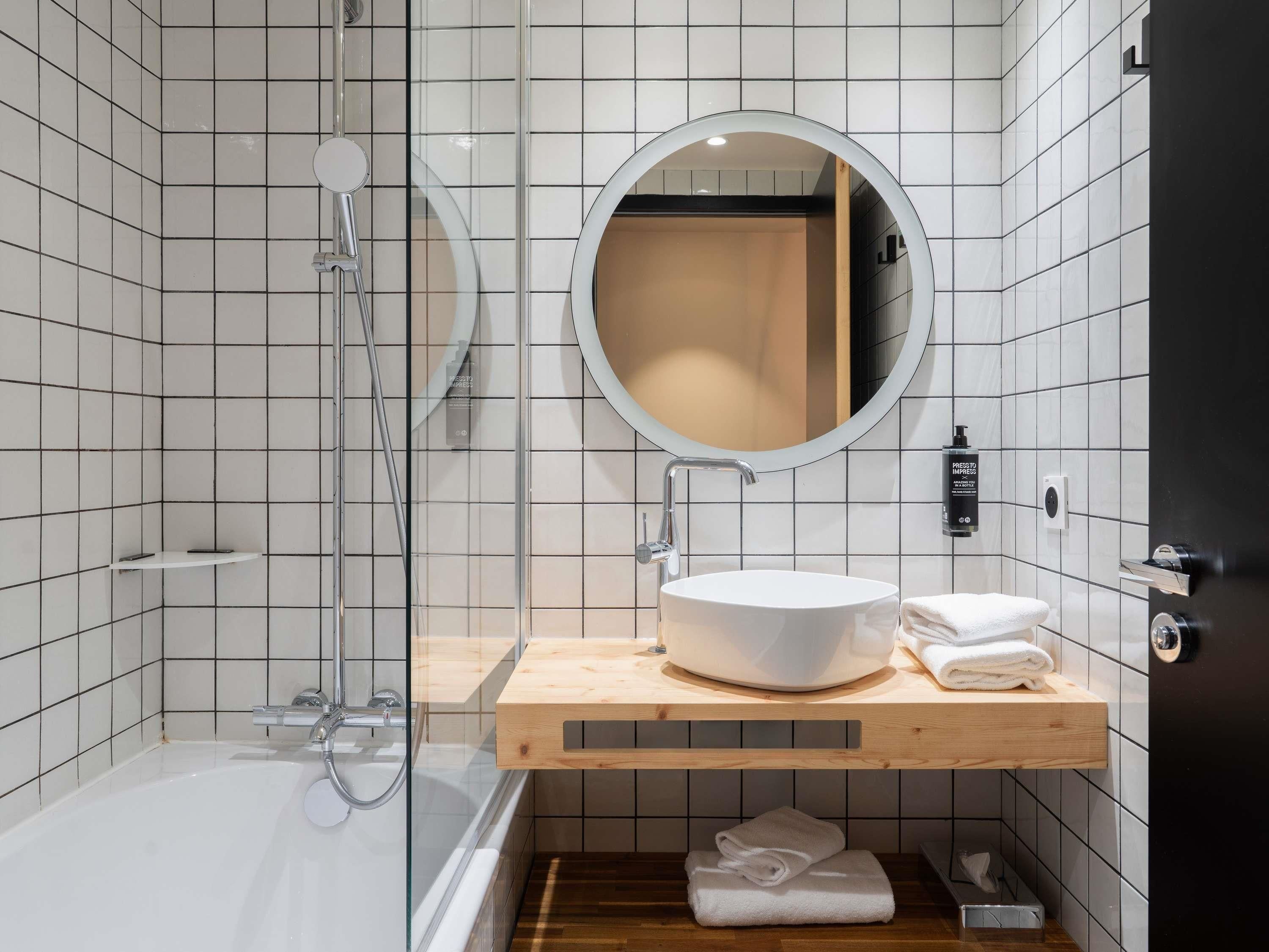 Un baño moderno con ducha de vidrio, un espejo redondo, un lavamanos blanco y un mueble de tocador de madera.